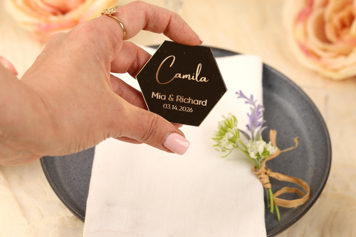 A hand holding a black hexagonal card with the name 'Camila' and the date '03.14.2026' printed on it, placed on a white napkin with a floral arrangement.