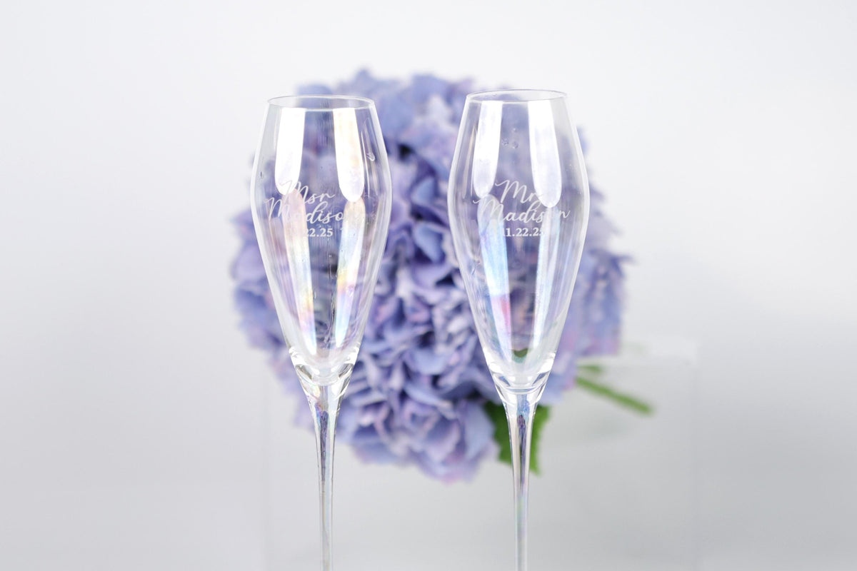Two champagne glasses with personalized messages stand in front of a bouquet of purple flowers.