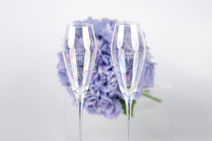 Two champagne glasses with personalized messages stand in front of a bouquet of purple flowers.
