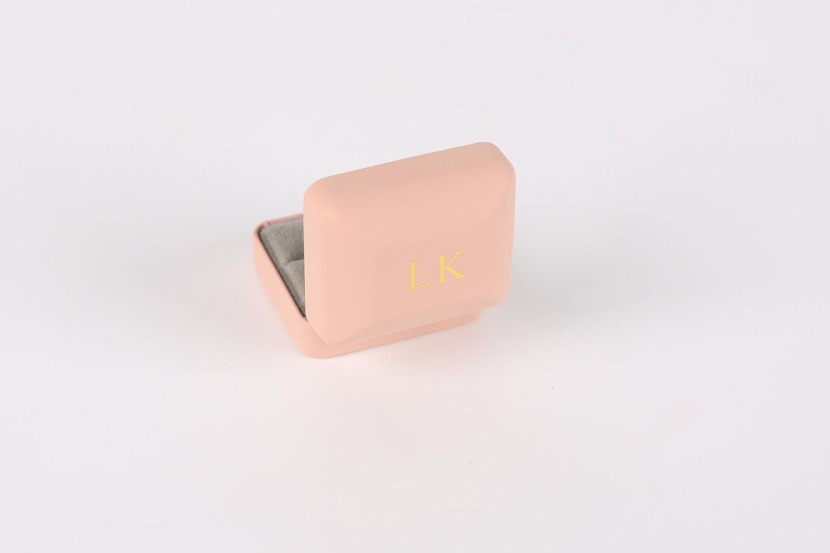 A pink jewelry box with a gold-colored interior, featuring a gold-colored letter "LK" on the front.
