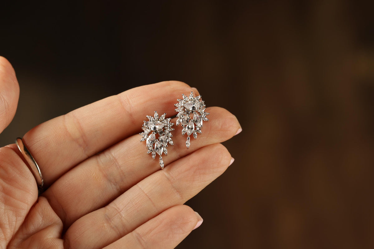 A hand holding a pair of elegant diamond earrings against a blurred background.