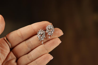 A hand holding a pair of elegant diamond earrings against a blurred background.