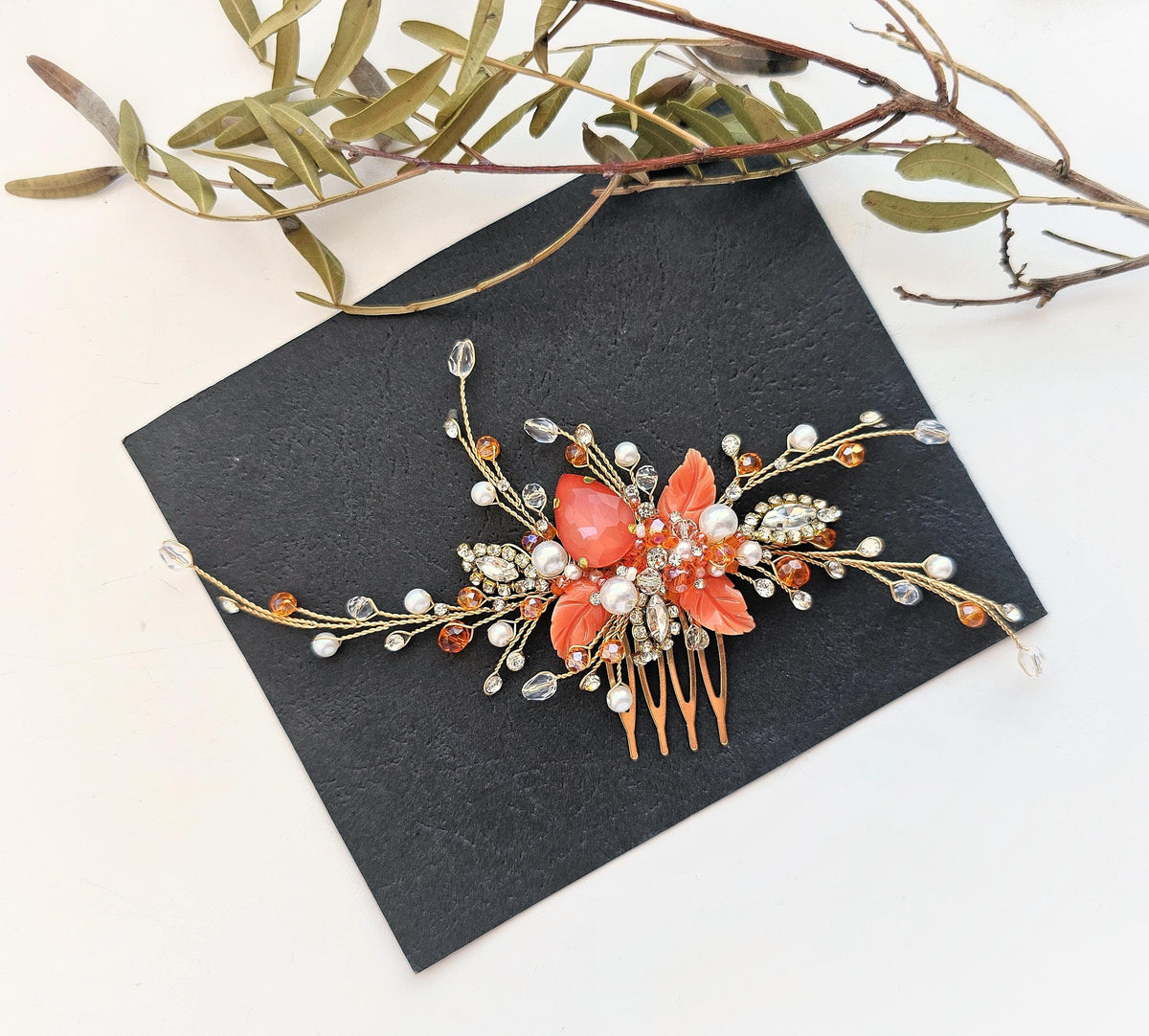 A decorative hair accessory with a floral design and sparkling embellishments, including a large red flower and numerous small pearls, is placed on a black surface alongside some green leaves.