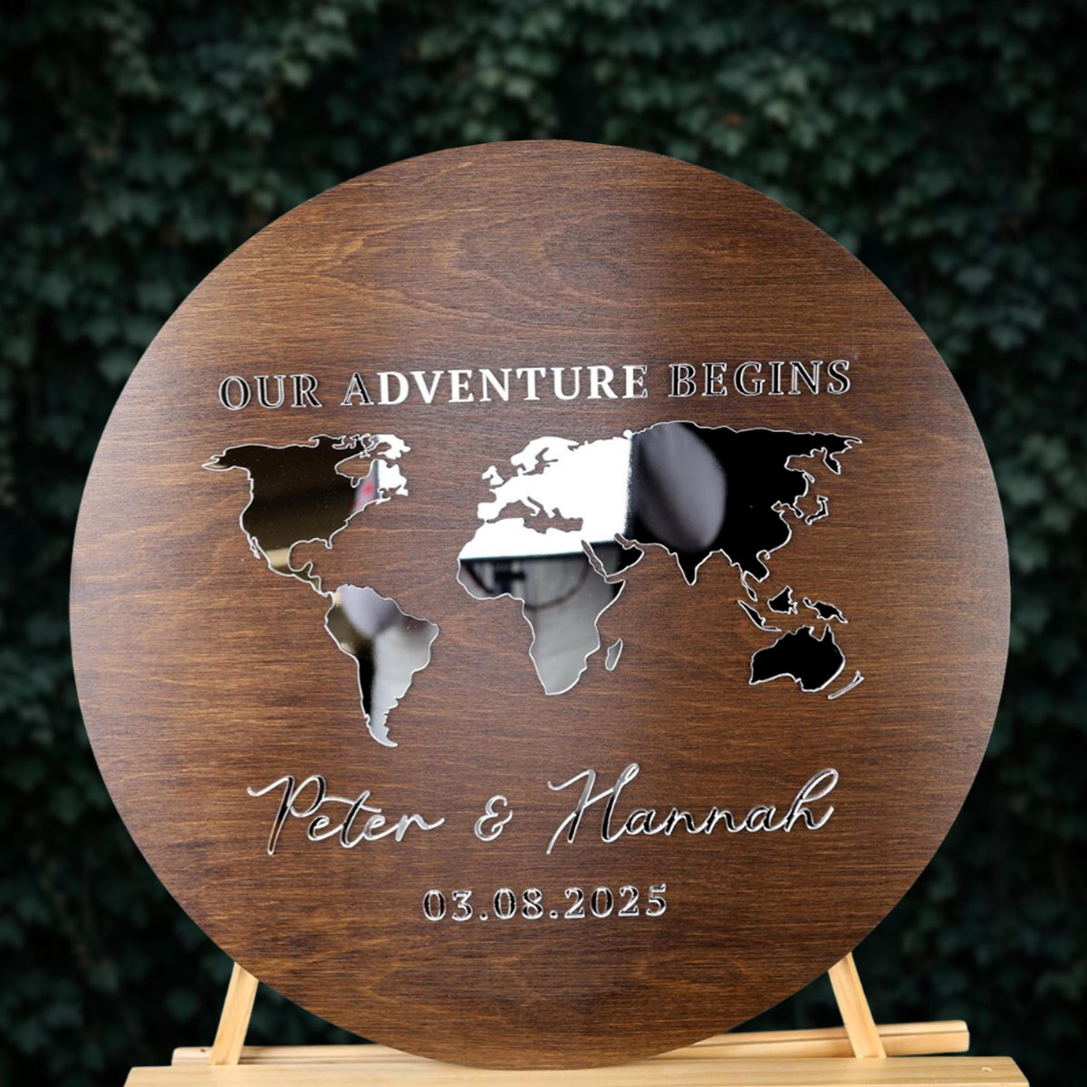 A wooden sign with a world map and the text "OUR ADVENTURE BEGINS" and "Peter & Hannah 03.08.2025" written on it.