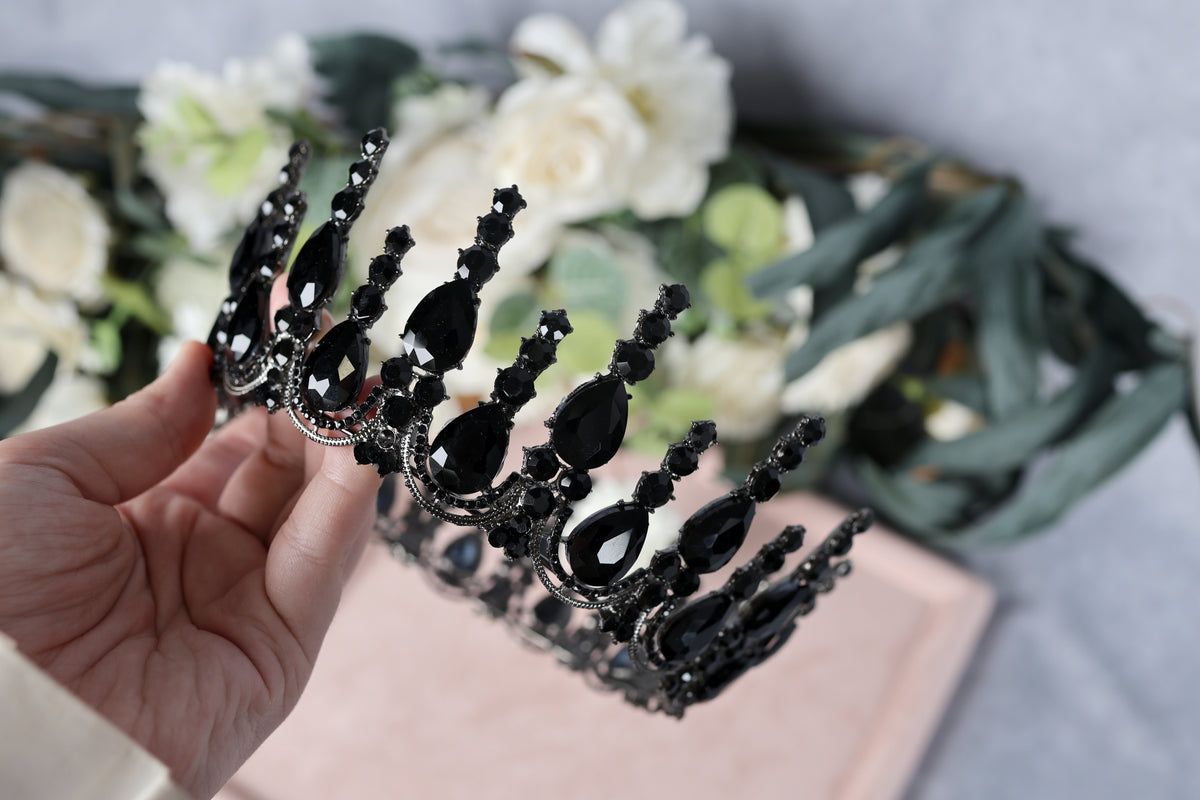 A hand is holding a black ornate crown adorned with black gemstones, with a blurred background featuring white flowers.