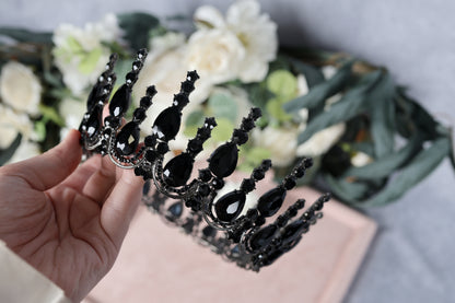 A hand is holding a black ornate crown adorned with black gemstones, with a blurred background featuring white flowers.