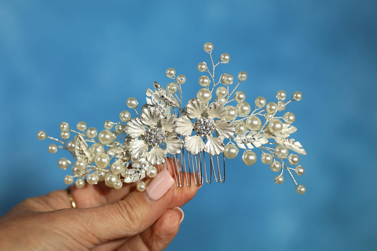 A hand holding a decorative hair accessory with white pearls and silver flowers against a blue background.