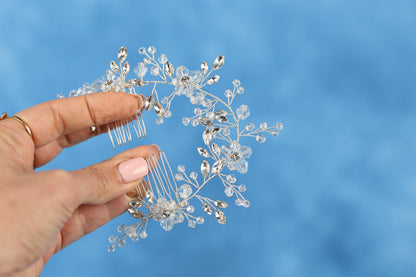 A hand holding a decorative hair accessory with clear crystal or gemstone embellishments against a blue background.