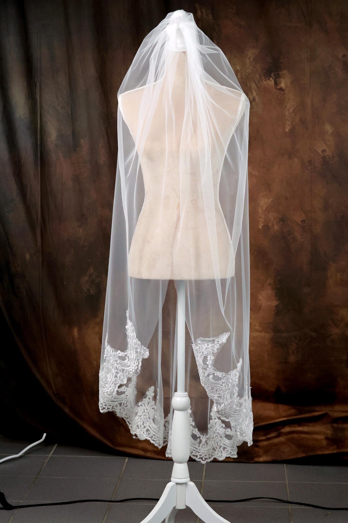A white mannequin wearing a white veil with intricate lace details, placed against a brown background.