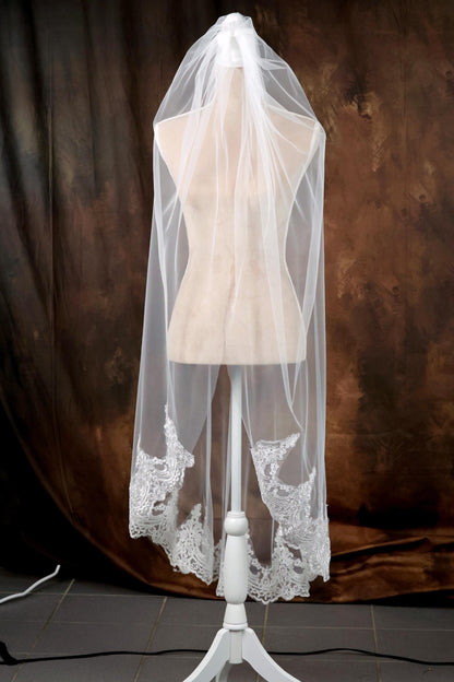 A white mannequin wearing a white veil with intricate lace details, placed against a brown background.
