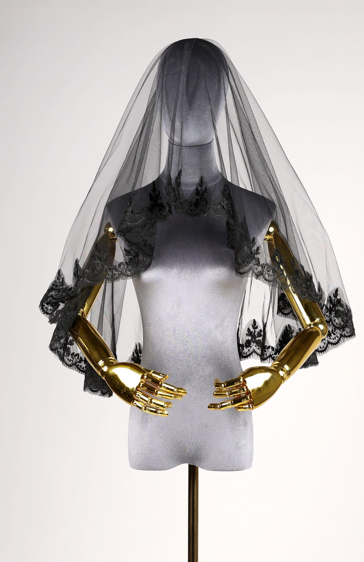 A mannequin wearing a black veil with lace trim and gold-colored hands, displayed against a plain white background.