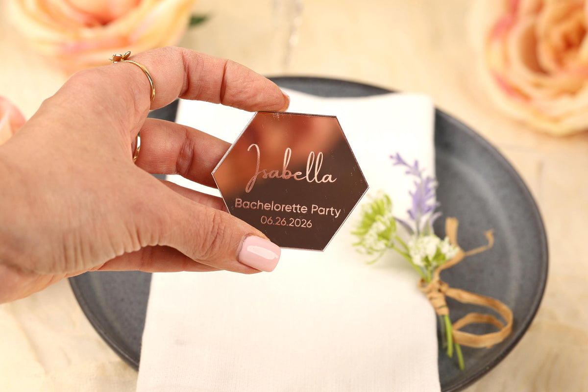 A hand holding a hexagonal metallic tag with the name "Isabella" and the date "Bachelorette Party" printed on it, with a floral arrangement and a plate visible in the background.