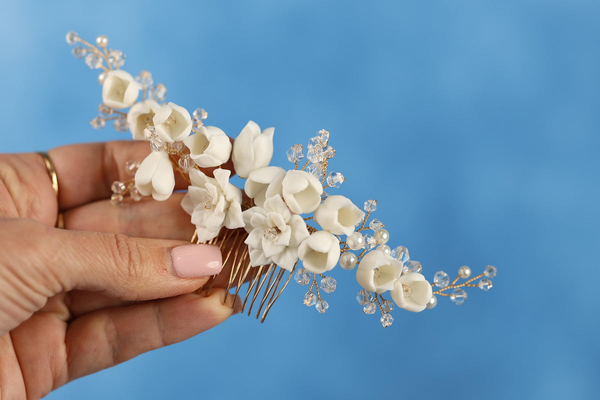 A hand holding a decorative hair clip with white flowers and clear beads against a blue background.
