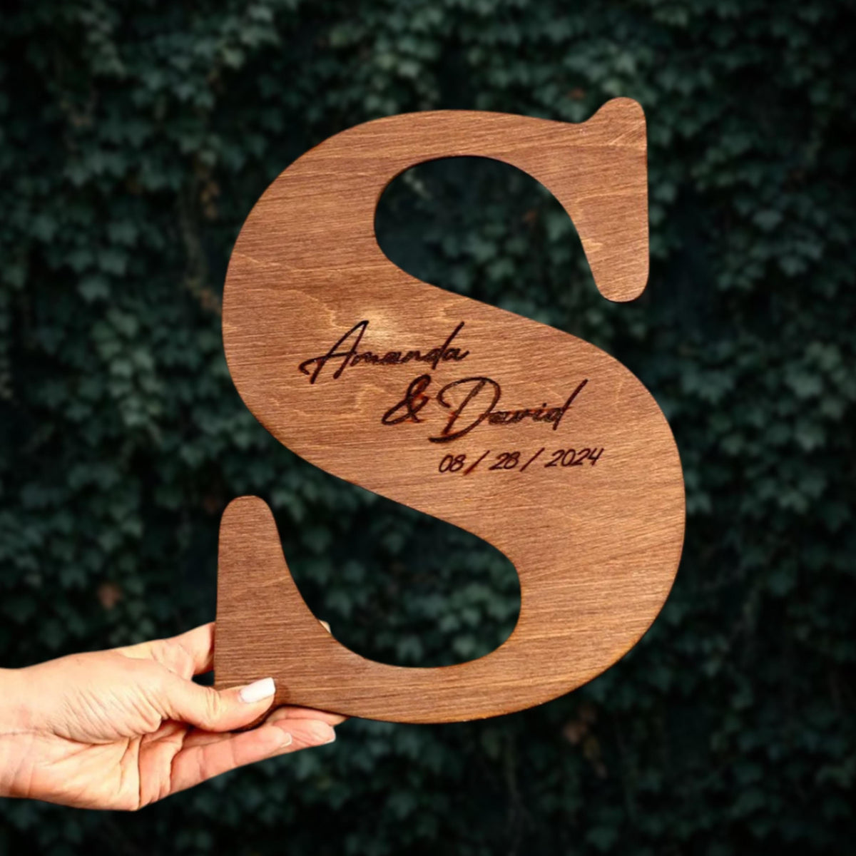 A hand holding a wooden letter 'S' with the names 'Amanda & David' and the date '08/29/2024' engraved on it.