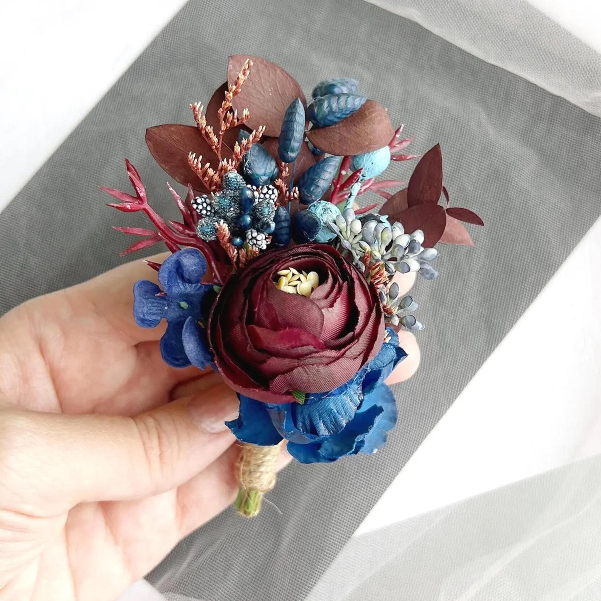 A hand is holding a colorful floral boutonniere made of dried flowers, including a large red rose, blue flowers, and other natural elements.