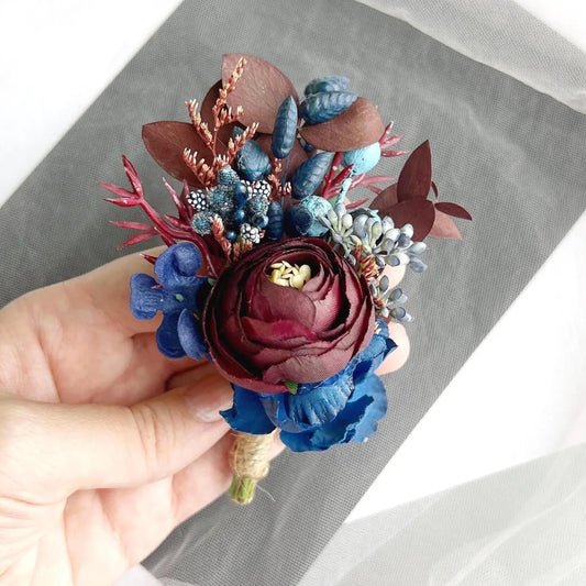 A hand is holding a colorful floral boutonniere made of dried flowers, including a large red rose, blue flowers, and other natural elements.