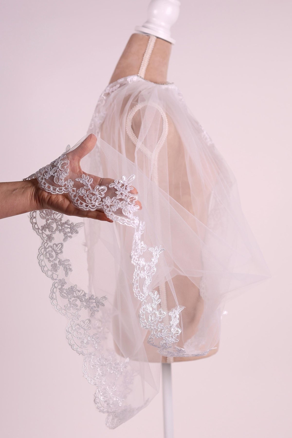 A hand is holding a delicate white lace veil with intricate floral patterns, which is displayed on a mannequin.