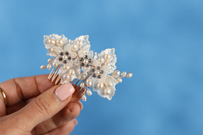 A hand holding a decorative hair accessory with white flowers and pearls against a blue background.