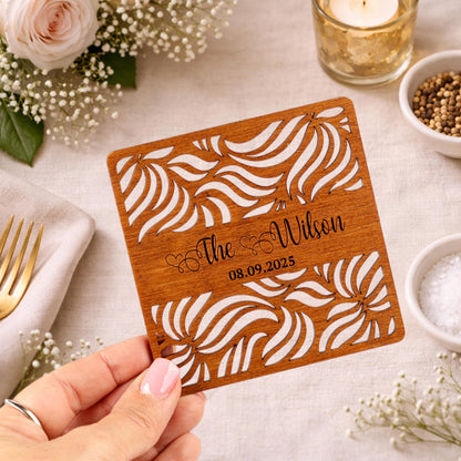 A hand holding a wooden card with a floral design and text on it, surrounded by other decorative items such as flowers, a candle, and a bowl of spices.
