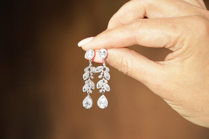 A hand holding a pair of diamond earrings with intricate leaf designs.