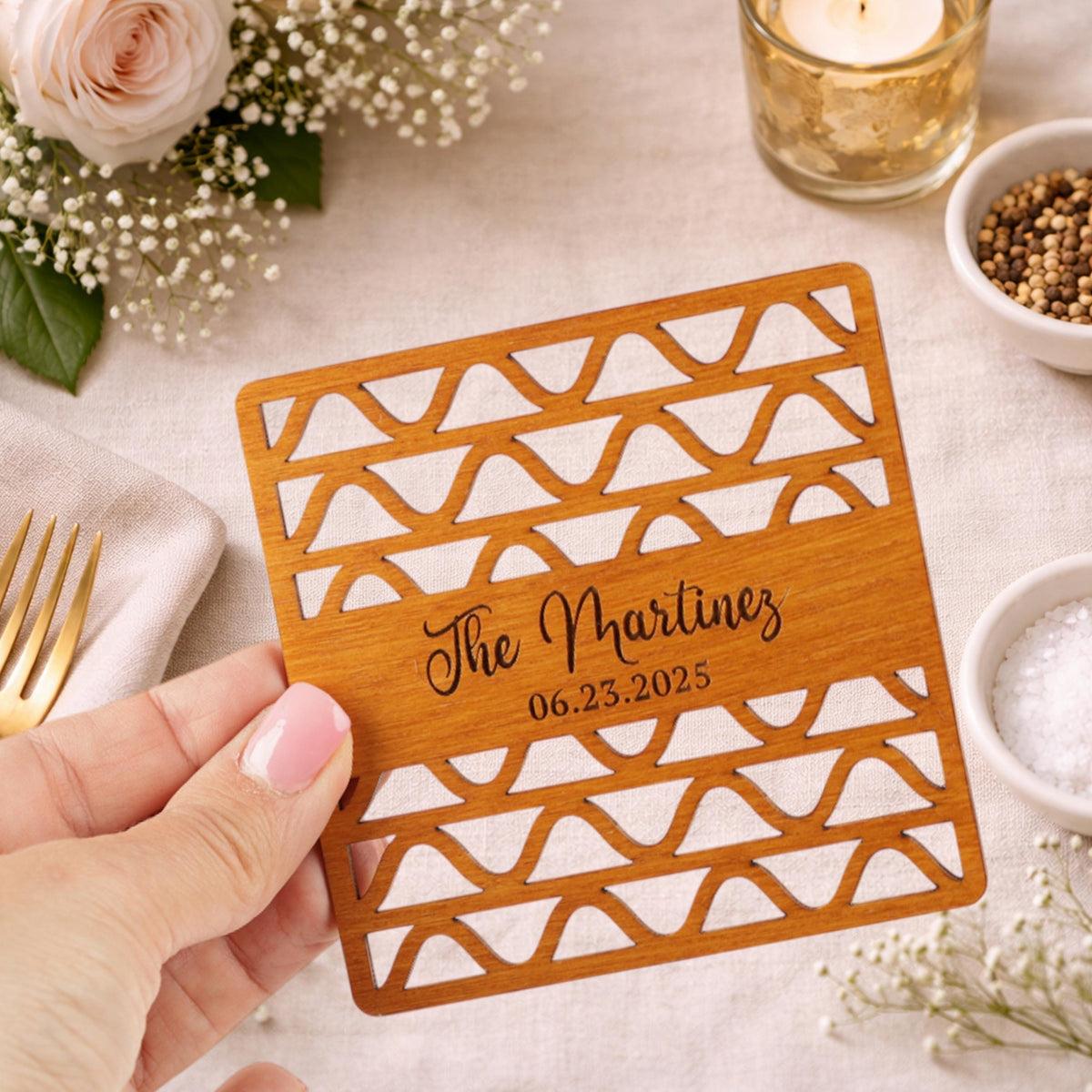 A hand holding a wooden coaster with text and a date printed on it, surrounded by other items such as a rose, a candle, and a bowl of spices.