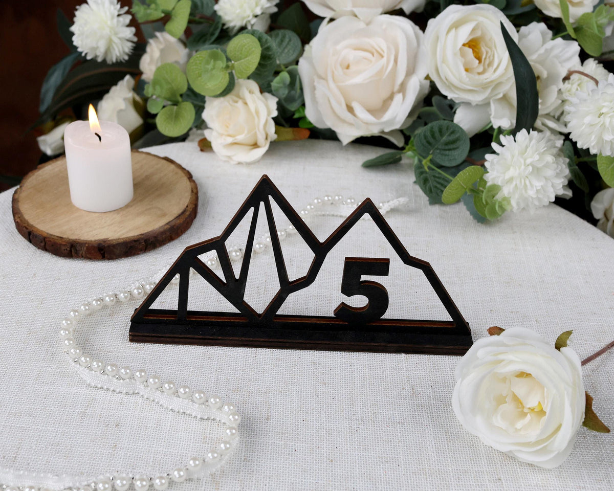 a wooden mountain-shaped sign with the number 5 on it, surrounded by white flowers and a lit candle on a table.