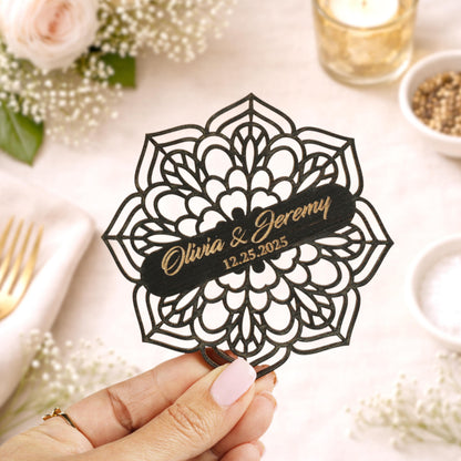 A hand holding a decorative paper with the names 'Olivia & Jeremy' and the date '12.25.25' on it, surrounded by floral decorations and other wedding-related items.