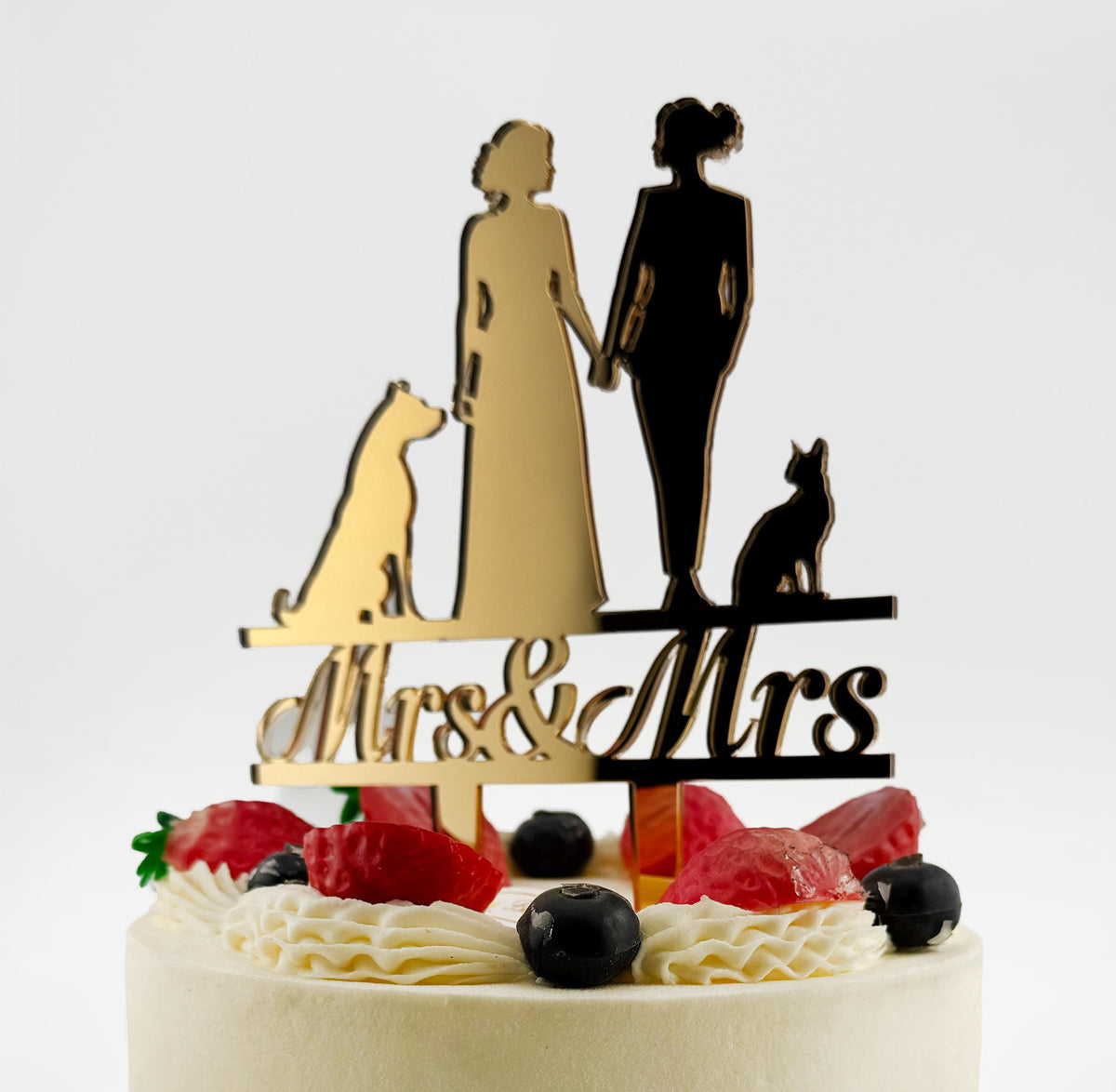 A wedding cake with a gold-colored topper featuring silhouettes of a bride and groom holding hands, with the words "Mrs & Mrs" written below.