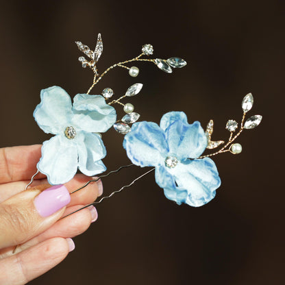 A hand holding a pair of decorative hair clips with blue and white flowers and sparkling rhinestones.