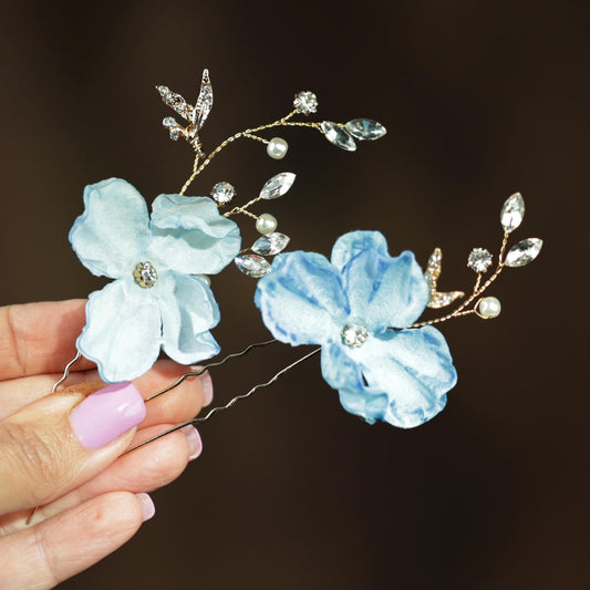 A hand holding a pair of decorative hair clips with blue and white flowers and sparkling rhinestones.