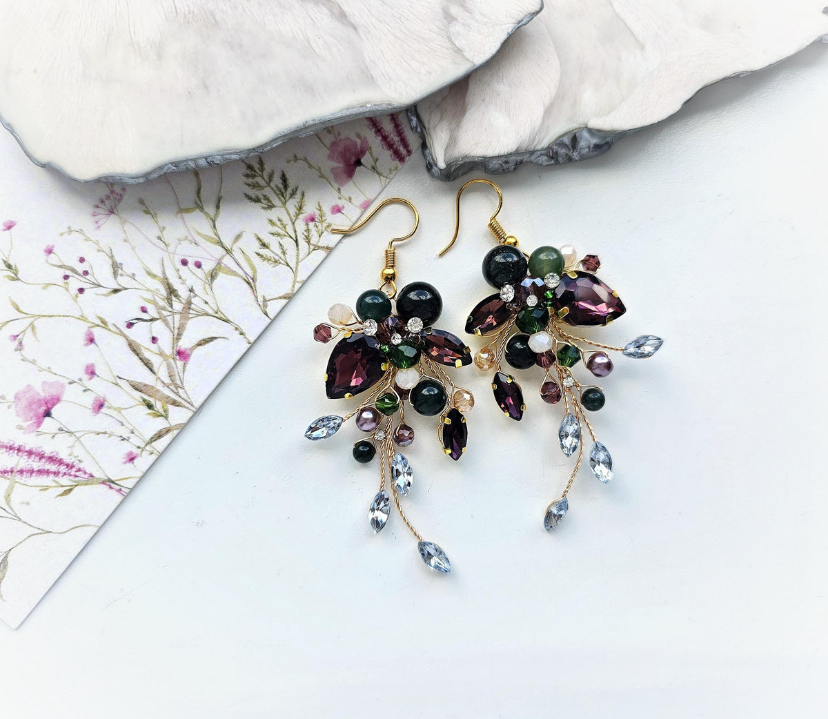 A pair of ornate earrings with a floral design and various colorful gemstones, including purple, green, and blue stones, are displayed on a white surface.