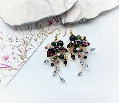 A pair of ornate earrings with a floral design and various colorful gemstones, including purple, green, and blue stones, are displayed on a white surface.