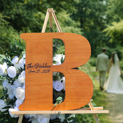 A wooden letter 'B' with the text "The Bishkos June 21, 2024" engraved on it, standing on a wooden stand surrounded by white flowers. In the background, there is a blurred image of a couple walking.