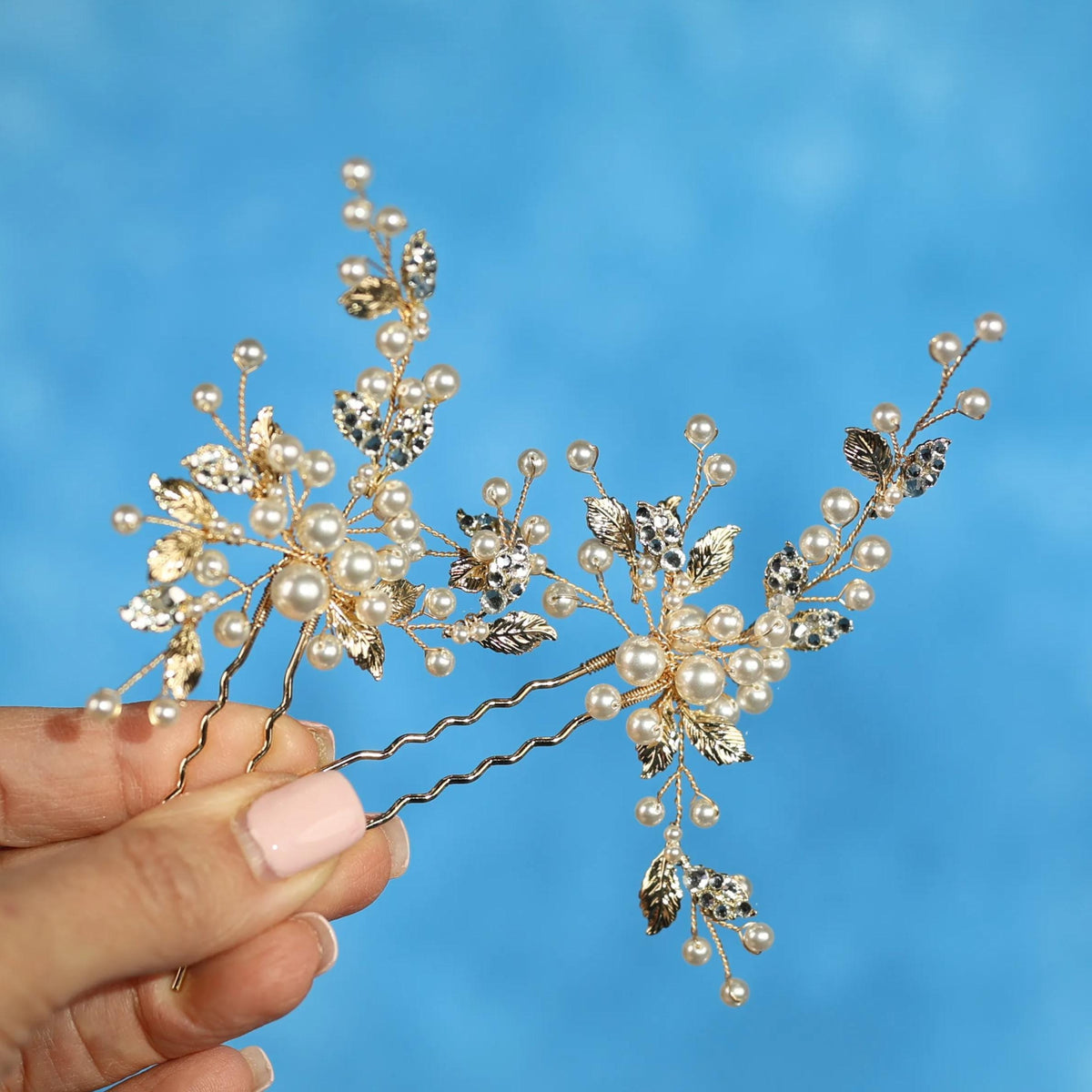 A hand holding a decorative hair clip with small pearls and leaves on a blue background.