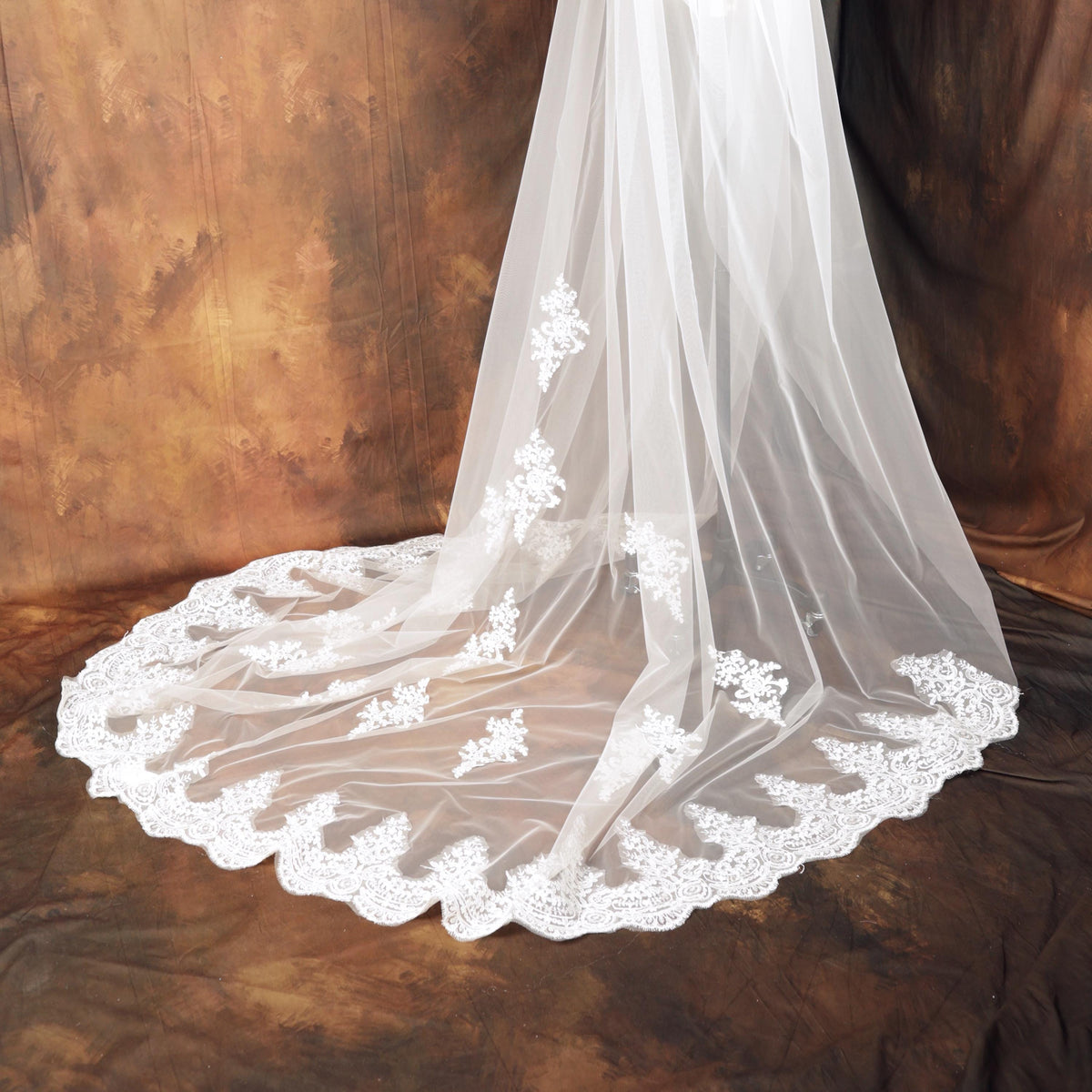 A white wedding veil with a delicate lace trim, featuring a floral pattern, is displayed against a brown background.
