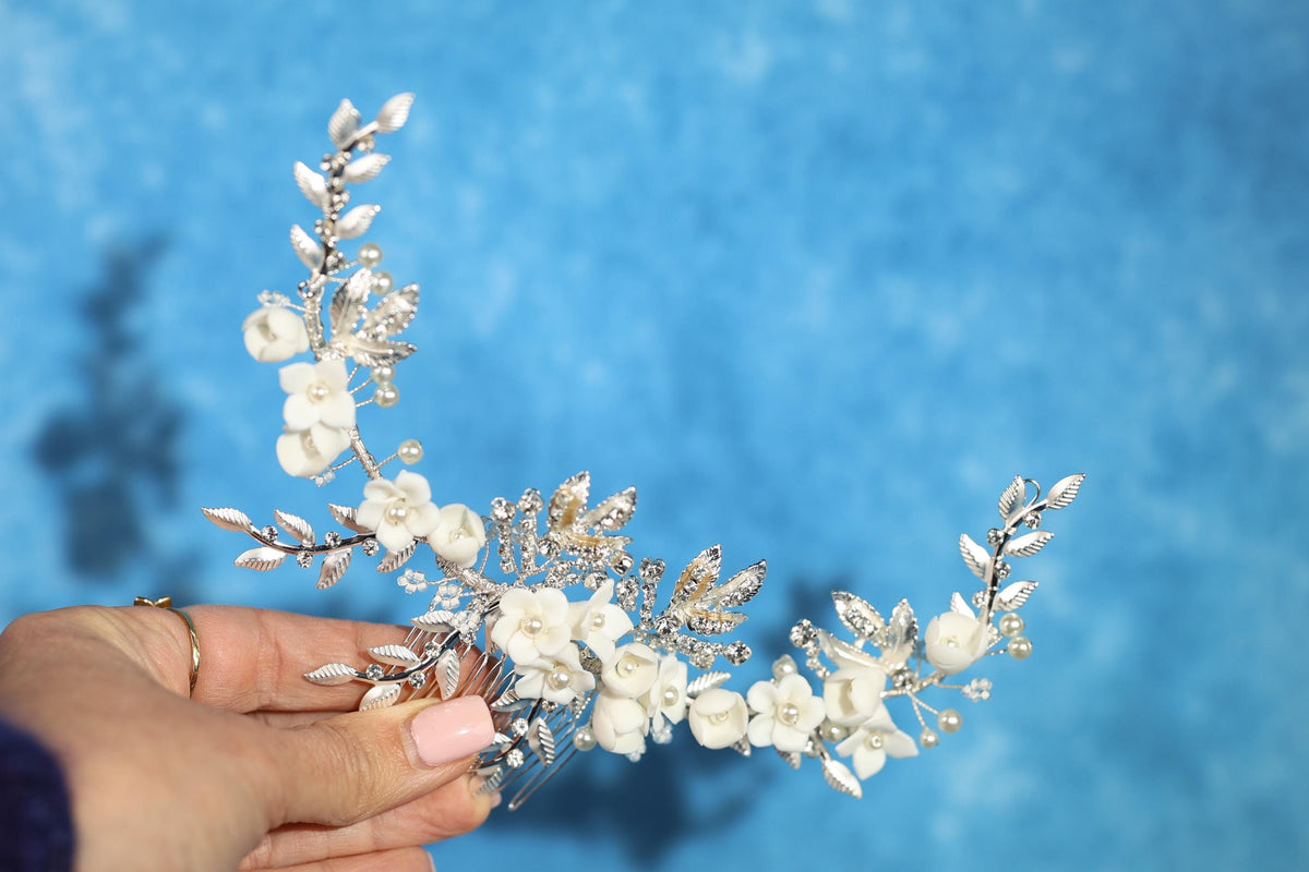 A hand holding a decorative hair accessory with white flowers and leaves against a blue background.