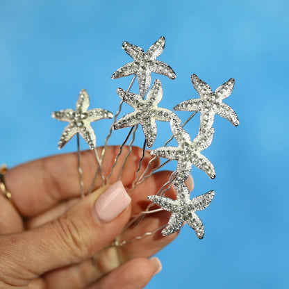 A hand holding a silver hair clip with five star-shaped embellishments against a blue background.