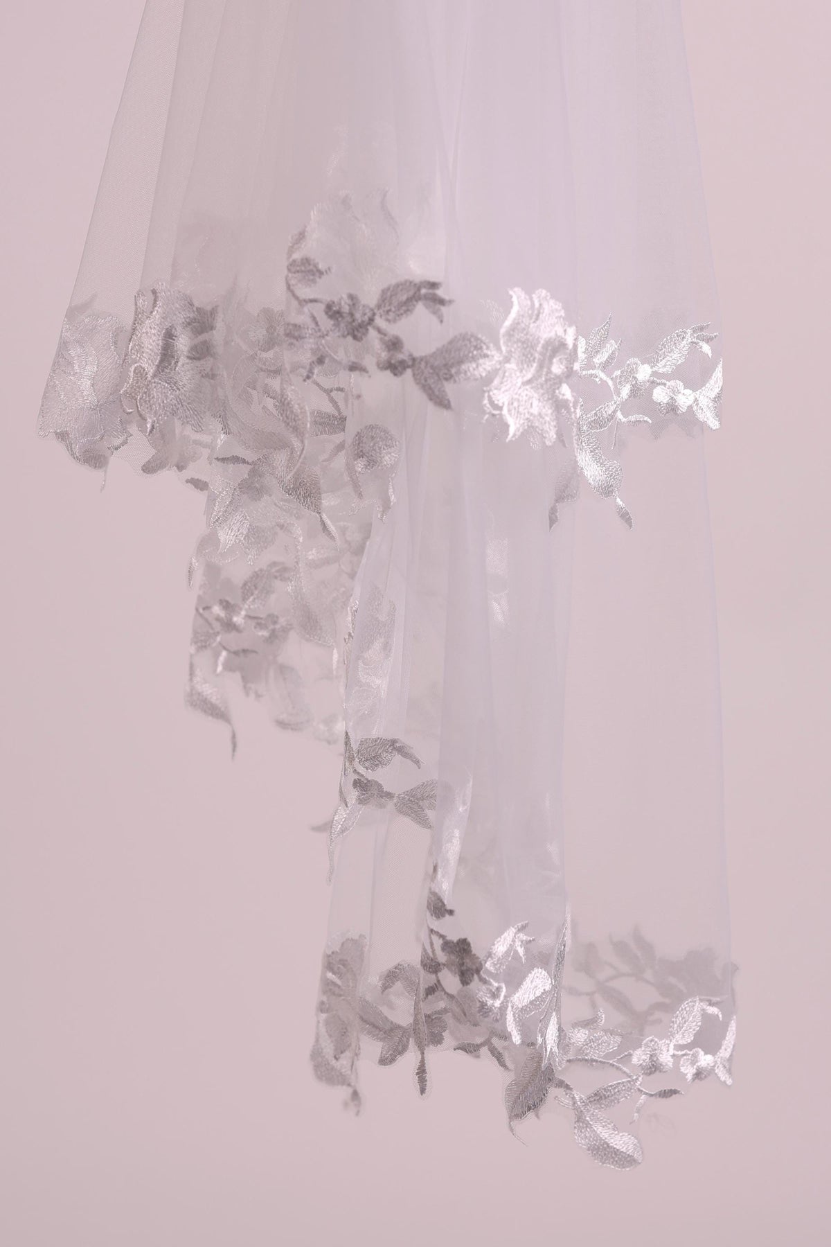 A delicate white veil with intricate silver embroidery, hanging against a soft pink background.