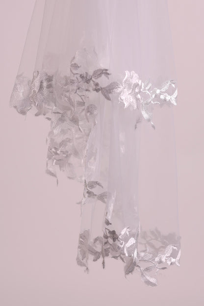 A delicate white veil with intricate silver embroidery, hanging against a soft pink background.