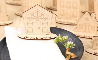 a wooden house-shaped place card holder with a nameplate, placed on a table with a plate of flowers and a small bouquet.