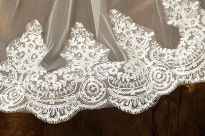 A close-up view of a delicate, intricately designed white lace fabric, featuring a floral pattern and a scalloped edge.