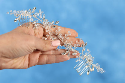 A hand holding a decorative hair accessory with clear crystal or crystal-like elements against a blue background.