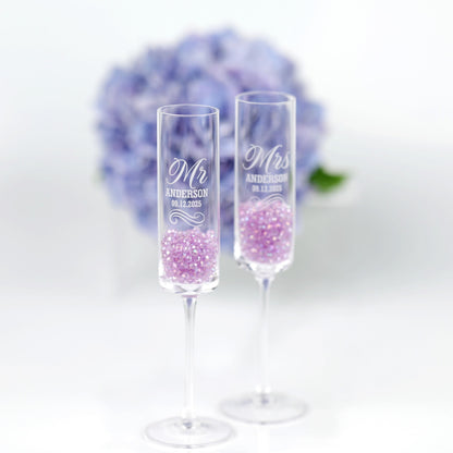 two clear champagne glasses filled with purple glitter, placed side by side against a blurred background of a bouquet of blue flowers.