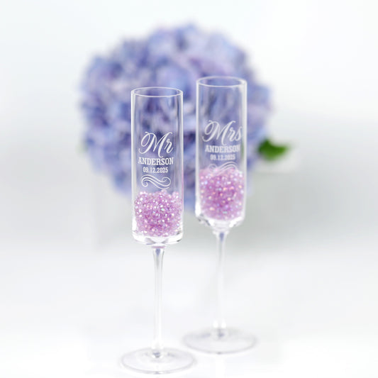 two clear champagne glasses filled with purple glitter, placed side by side against a blurred background of a bouquet of blue flowers.