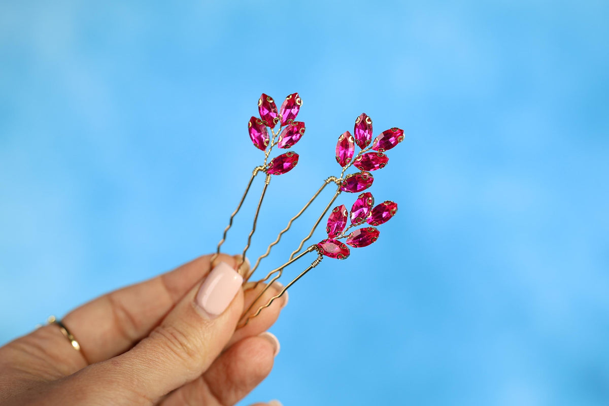 A hand holding a pair of gold hair clips with red gemstone branches on a blue background.