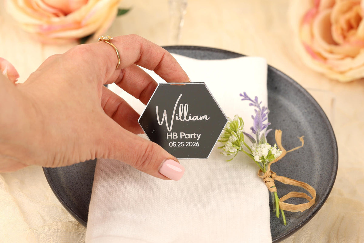 A hand holding a hexagonal black card with the name "William" and the date "05.25.2026" on it, placed on a white napkin with a floral decoration.