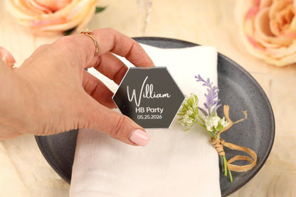 A hand holding a hexagonal black card with the name "William" and the date "05.25.2026" on it, placed on a white napkin with a floral decoration.