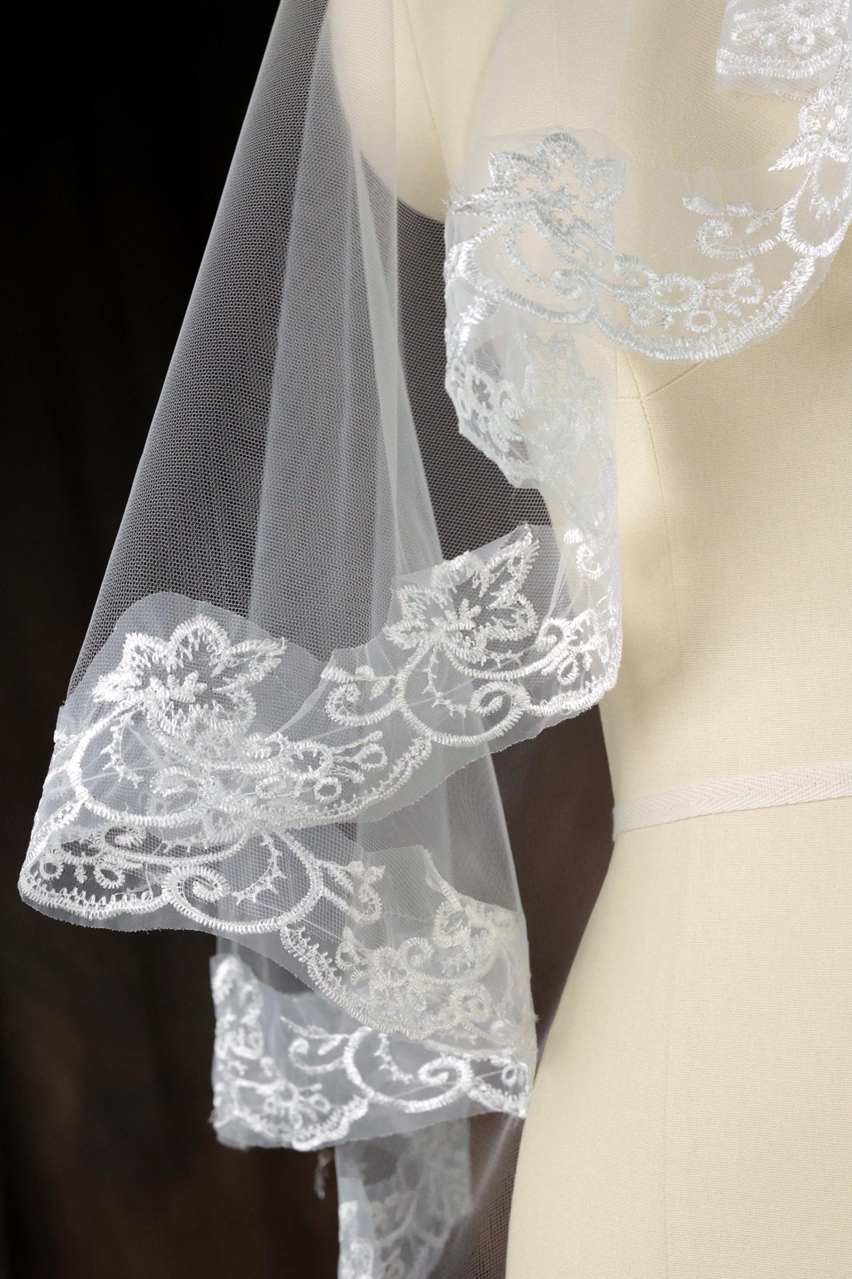 A close-up view of a delicate white lace veil with intricate floral embroidery, displayed on a mannequin.