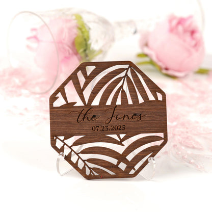 A wooden coaster with a palm leaf design and the name "The Jones" printed on it, placed on a white surface with a pink rose in the background.
