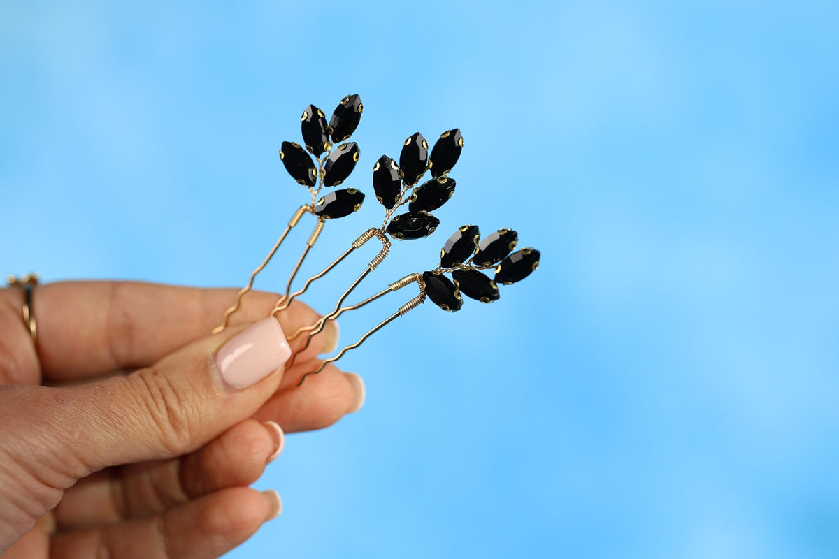 A hand holding three gold-colored hair clips with black gemstone leaves attached to them against a blue background.